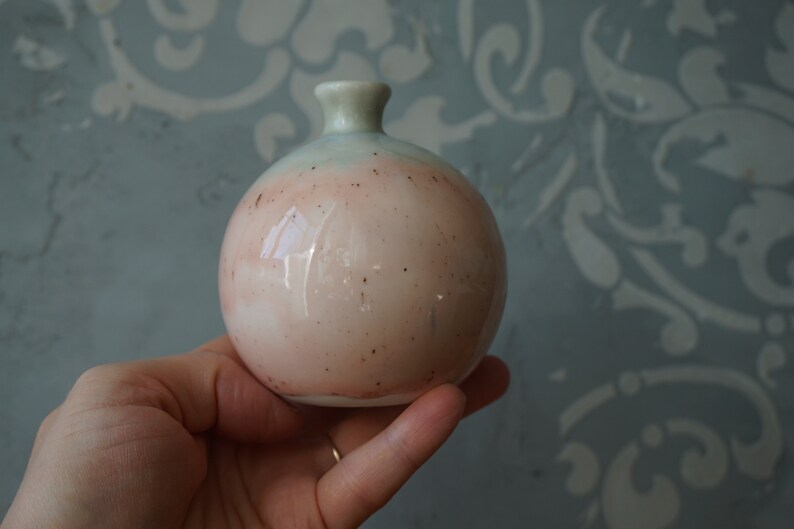 K&ouml;nnte beinhalten: Eine kleine, runde Keramikvase mit hellblauer und rosafarbener Glasur. Die Vase hat eine gesprenkelte Oberfl&auml;che und eine leicht abgerundete &Ouml;ffnung.