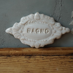 Può includere: Un'insegna da bagno in ceramica bianca con la scritta "BAGNO" in rilievo al centro. L'insegna ha un bordo ornato e decorativo ed è posta contro un muro grigio e una superficie in legno.