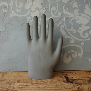 May include: A gray plaster hand sculpture with five fingers. The sculpture is standing on a wooden surface.