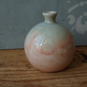 K&ouml;nnte beinhalten: Eine kleine, runde Keramikvase mit hellgr&uuml;ner und rosafarbener Glasur. Die Vase hat eine gesprenkelte Oberfl&auml;che und eine schmale &Ouml;ffnung oben.