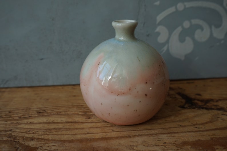 K&ouml;nnte beinhalten: Eine kleine, runde Keramikvase mit hellgr&uuml;ner und rosafarbener Glasur. Die Vase hat eine gesprenkelte Oberfl&auml;che und eine schmale &Ouml;ffnung oben.