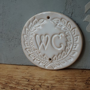 May include: White ceramic round sign with a wreath of leaves around the letters "WC".