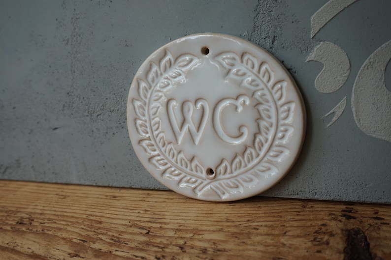 May include: White ceramic round sign with a wreath of leaves around the letters "WC".