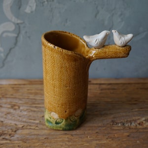 May include: A ceramic vase with a textured, woven-like pattern in shades of yellow and brown. Two small white bird figurines perch on a branch extending from the vase. The base has green and white accents.