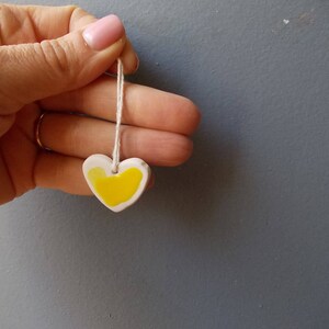 May include: A white ceramic heart pendant with a yellow painted design, hanging from a white string.