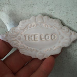 May include: A white ceramic plaque with the text "THE LOO" in raised letters. The plaque has an ornate, decorative border with a light beige accent. The plaque is diamond-shaped and appears to be a bathroom sign.