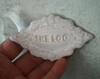 May include: A white ceramic plaque with the text "THE LOO" in raised letters. The plaque has an ornate, decorative border with a light beige accent. The plaque is diamond-shaped and appears to be a bathroom sign.