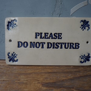Puede incluir: Un letrero de cerámica blanca con detalles florales azules y el texto "PLEASE DO NOT DISTURB".