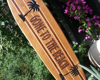Könnte beinhalten: Ein Holzsurfboard mit braunem und weißem Streifenmuster. Das Surfboard trägt den Text "Gone to the Beach" und zwei Palmen.