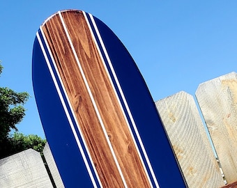 Könnte beinhalten: Ein blaues und braunes Surfbrett mit weißen Streifen und einem weißen Hibiskus-Blütenmuster. Das Surfbrett lehnt an einem Holzzaun.
