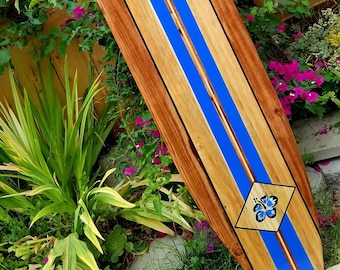 Könnte beinhalten: Ein Holzsurfboard mit blauen Streifen und einem blauen Blumendesign. Das Surfboard lehnt an einem Holzzaun.