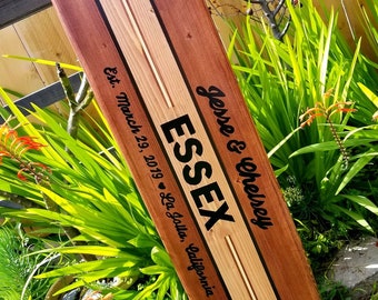 Puede incluir: Una tabla de surf de madera con un diseño a rayas blanco y negro. La tabla de surf está grabada con el texto "Jesse & Chelsey ESSEX Est. March 29, 2019 - La Jolla, California".
