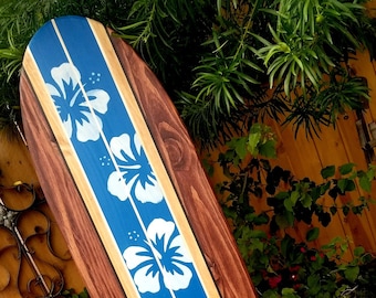 May include: A wooden surfboard painted with a blue and white hibiscus flower design. The surfboard is brown with a light brown stripe down the center.