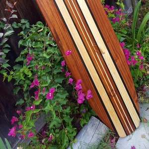 May include: A wooden surfboard with a unique design featuring alternating stripes of light and dark wood. The surfboard is decorated with black stripes and a sun design.