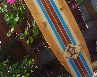 May include: A wooden surfboard with blue and brown stripes. The surfboard has a diamond-shaped design with a turtle in the center.