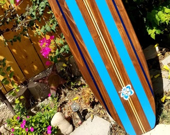 Könnte beinhalten: Ein Holzsurfboard, das mit blauen und weißen Streifen bemalt ist. Das Surfboard hat eine braune Holzmaserung und eine blaue Hibiskusblüte, die auf die Unterseite gemalt ist.