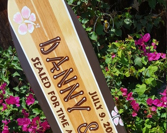 Puede incluir: Una tabla de surf de madera pintada con los nombres "Danny & Reui" y la fecha "July 9, 2016". La tabla de surf está decorada con flores de hibisco rosas y azules. El texto "Sealed for time and all eternity" está escrito en la tabla de surf.