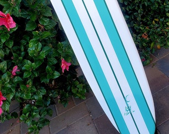 Könnte beinhalten: Ein weißes Surfbrett mit türkisfarbenen Streifen und einem türkisfarbenen Hai-Flossen-Design. Das Surfbrett lehnt an einem grünen Busch.