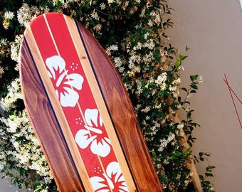 May include: A wooden surfboard with a red and white stripe design and white hibiscus flowers painted on it.