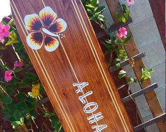 May include: A wooden surfboard with a brown stain and white stripes. The surfboard has two painted hibiscus flowers and the word "ALOHA" written in white.
