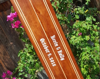 Puede incluir: Una tabla de surf de madera pintada de marrón con rayas blancas. La tabla de surf está personalizada con el texto "Kevin & Kelly October 9, 2016".