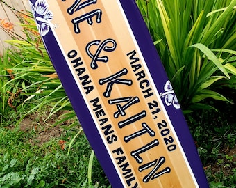 Puede incluir: Una tabla de surf de madera pintada con rayas moradas y marrones. La tabla de surf tiene el texto "Shane & Kaitlin Johns" en blanco con contorno negro. El texto "March 21, 2020" también está en la tabla de surf. El texto "Ohana means family" está escrito en la tabla de surf.