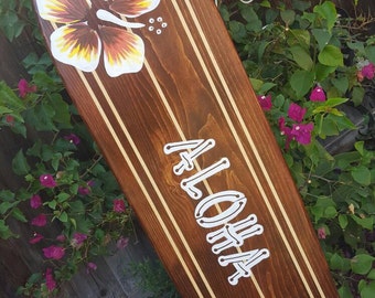 May include: A brown wooden surfboard with white stripes and two yellow hibiscus flowers painted on it. The word "ALOHA" is painted in white on the surfboard.