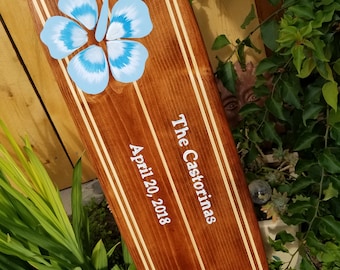 May include: A wooden surfboard with two blue hibiscus flowers painted on it. The surfboard is decorated with white stripes and the text "The Castorinas April 20, 2018".