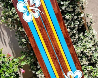 May include: A wooden surfboard painted with blue and yellow stripes and two white hibiscus flowers. The surfboard is leaning against a wall with green plants.