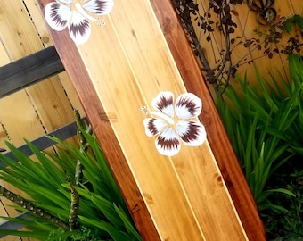 May include: Wooden surfboard with a light brown center and dark brown edges. The surfboard is decorated with three white and brown hibiscus flower decals. The board is leaning against a wooden fence and surrounded by greenery.
