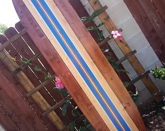 May include: A wooden surfboard with blue, white, and brown stripes. The surfboard is leaning against a wooden fence and has the text "San Diego Surf Art" on the bottom.