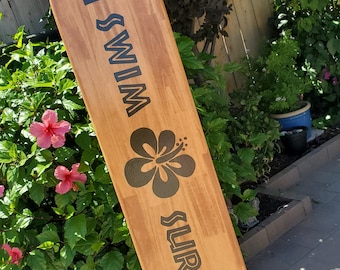 Könnte beinhalten: Ein schwarzes, bemaltes Holzsurfboard mit den Worten "Swim Surf" und einem schwarzen Hibiskus-Blumenmotiv.