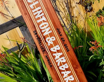 May include: A wooden surfboard painted with white stripes and two brown hibiscus flowers. The surfboard is decorated with the text "Clinton & Barbara" and "July 27, 2015 Seychelles, Africa".