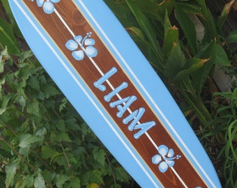 Könnte beinhalten: Ein blaues und braunes Surfbrett mit weißen Streifen und blauen Hibiskusblumen, die darauf gemalt sind. Der Name "LIAM" ist in Weiß auf das Surfbrett gemalt.