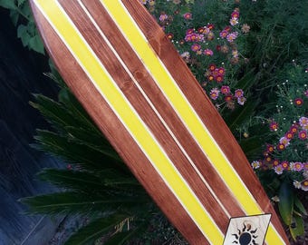 Könnte beinhalten: Ein Holzsurfboard mit gelben Streifen und einem schwarzen Sonnendesign am Heck. Das Surfboard steht auf einem Betonblock vor einem grünen Busch.