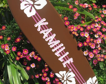 Könnte beinhalten: Ein braunes Surfbrett mit rosa Streifen und weißen Hibiskusblüten. Der Name "Kamille" ist in rosa auf dem Surfbrett geschrieben.