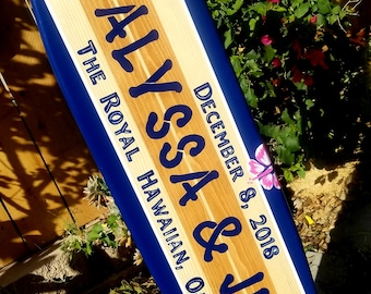 May include: A blue and wood surfboard with the text "Alyssa & Jeff" and "December 8, 2018 The Royal Hawaiian, Oahu, HI" in white lettering. The surfboard has pink hibiscus flowers painted on it.