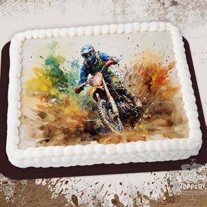 May include: A white square cake with a watercolor image of a motocross rider in a blue and white suit, riding a dirt bike through a muddy track. The cake is decorated with white frosting.