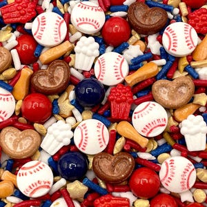 May include: A colorful mix of sprinkles featuring baseballs, bats, red spheres, blue and white rods, star-shaped sprinkles, and heart-shaped cookies. The sprinkles are in red, white, blue, gold, and brown colors.
