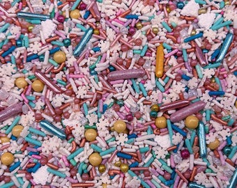 May include: A festive mix of edible sprinkles featuring white snowflake shapes, metallic gold and blue rod-shaped sprinkles, pink and turquoise jimmies, and small round sugar pearls in shades of gold, pink, blue, and white for cake decoration.