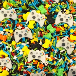 May include: A colorful assortment of sprinkles featuring white video game controllers, stars, and bone shapes. The sprinkles are in various colors, including blue, green, yellow, orange, and black, creating a playful and festive appearance. Ideal for decorating baked goods.