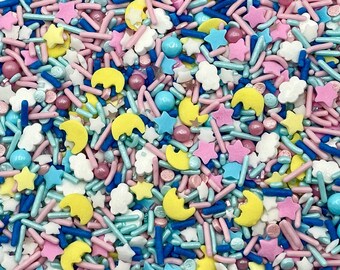 May include: A collection of edible cake sprinkles featuring a mix of yellow crescent moons, white clouds, pink and blue stars, and various rod-shaped sprinkles in shades of pink, light blue, dark blue, and white, along with small round spheres.