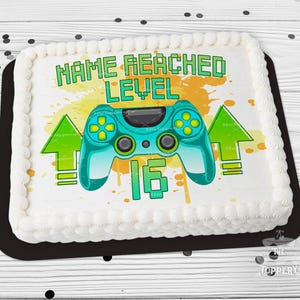 May include: A rectangular cake with white frosting and a black base. The cake features a teal video game controller graphic, green arrows, and the text "NAME REACHED LEVEL 16". The background has yellow and orange paint splatters.