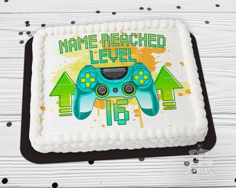 May include: A rectangular cake with white frosting and a black base. The cake features a teal video game controller graphic, green arrows, and the text "NAME REACHED LEVEL 16". The background has yellow and orange paint splatters.