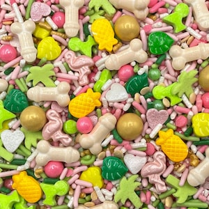 May include: A colorful mix of edible sprinkles featuring light beige phallic shapes, yellow pineapples, pink flamingos, green palm trees, green monstera leaves, white hearts, gold spheres, and pink round candies mixed with thin pink and green rods.
