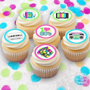 May include: Six cupcakes with white frosting and retro-themed edible toppers. Toppers feature a cassette tape, boombox, Rubik's Cube, television, and the phrase "I love the 80's". Colorful confetti dots the background.