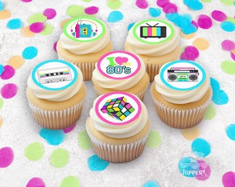 May include: Six cupcakes with white frosting and retro-themed edible toppers. Toppers feature a cassette tape, boombox, Rubik's Cube, television, and the phrase "I love the 80's". Colorful confetti dots the background.