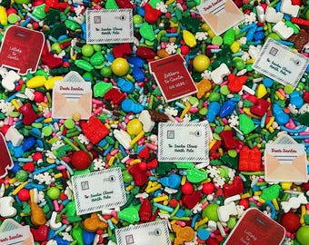 May include: A festive mix of edible sprinkles featuring red mailbox shapes with Letters to Santa text, white envelopes labeled To: Santa Claus North Pole, and Dear Santa envelopes, alongside red, green, blue, and yellow candy shapes and white snowflakes.