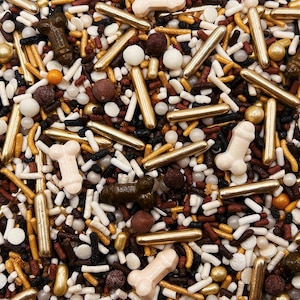 May include: A mix of edible sprinkles featuring metallic gold rods, small white and brown spheres, dark brown and cream-colored phallic-shaped pieces, and thin black, white, and brown cylindrical sprinkles scattered across a flat surface.