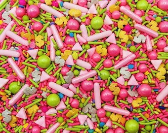 May include: A colorful assortment of cake sprinkles featuring pink rod-shaped sprinkles, bright green round candy balls, small yellow dollar sign shapes, silver dollar signs, and light pink triangles mixed with tiny blue and orange pieces.